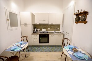 A compact kitchen with white cabinets, a built-in oven, and a dining area with blue table settings. - La Sicilia in un sogno (Syracuse)