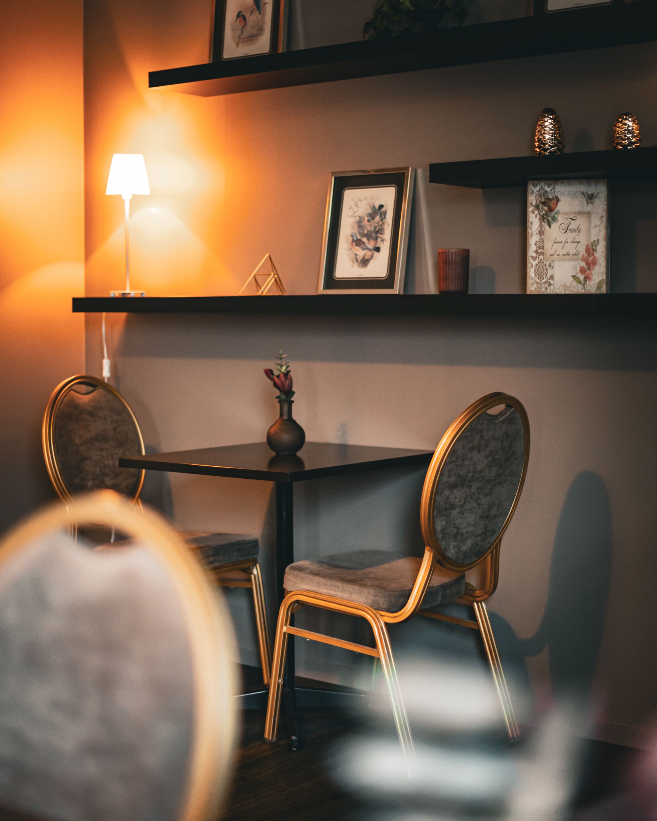 A dining area with a table and two chairs, a lamp, and framed artwork on shelves.