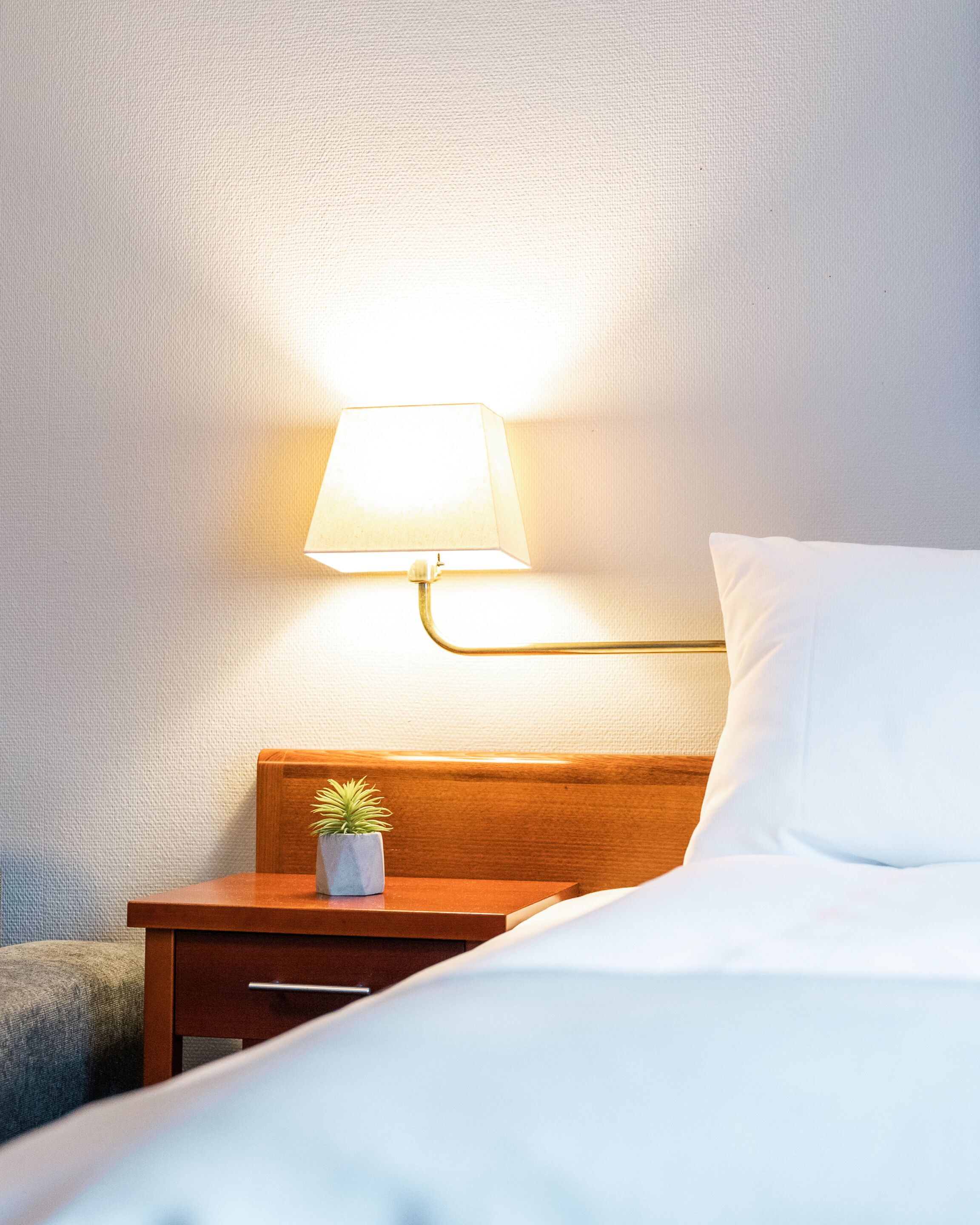 Una cama bien tendida con sábanas blancas, una mesita de noche de madera con una pequeña planta en maceta y una lámpara de pared.