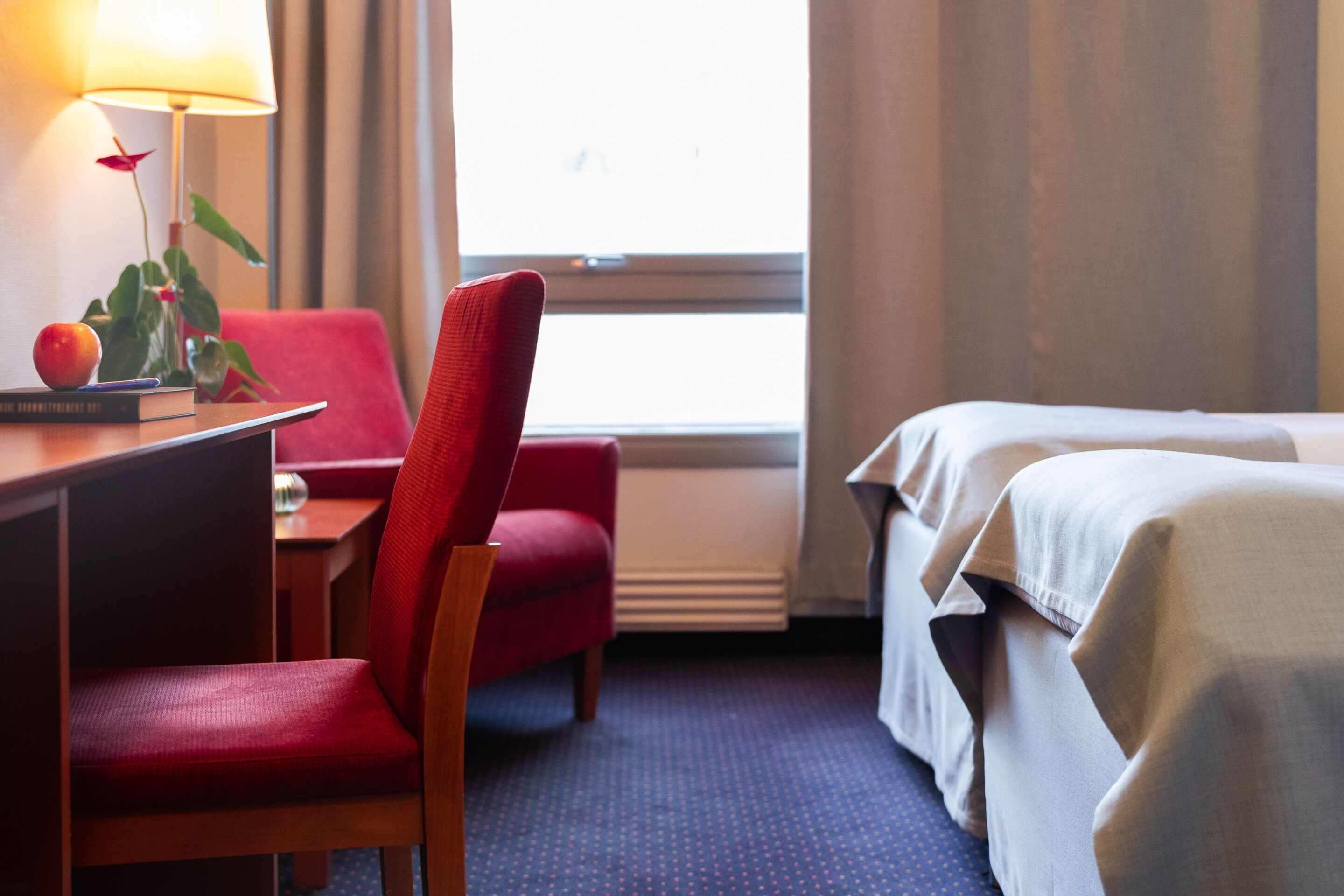 A hotel room with a desk, a red chair, a bed with white linens, a window with curtains, and a lamp on the desk.