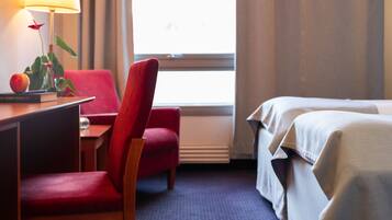 A hotel room with a desk, a red chair, a bed with white linens, a window with curtains, and a lamp on the desk.