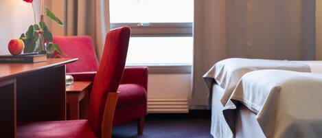 A hotel room with a desk, a red chair, a bed with white linens, a window with curtains, and a lamp on the desk.
