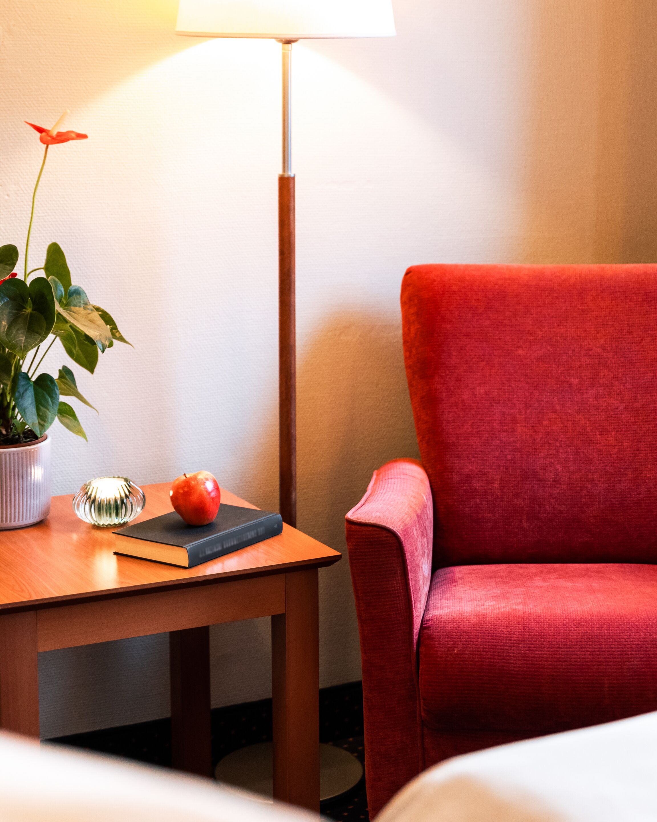 Habitación de hotel con una mesa de madera, un sillón rojo, una planta en maceta, un libro y una lámpara.