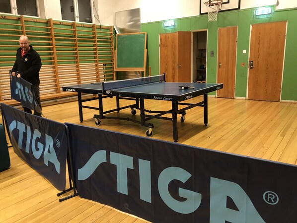 Une table de ping-pong ornée du logo de la marque « STIGA », un plancher en bois et un mur vert avec une porte en arrière-plan.