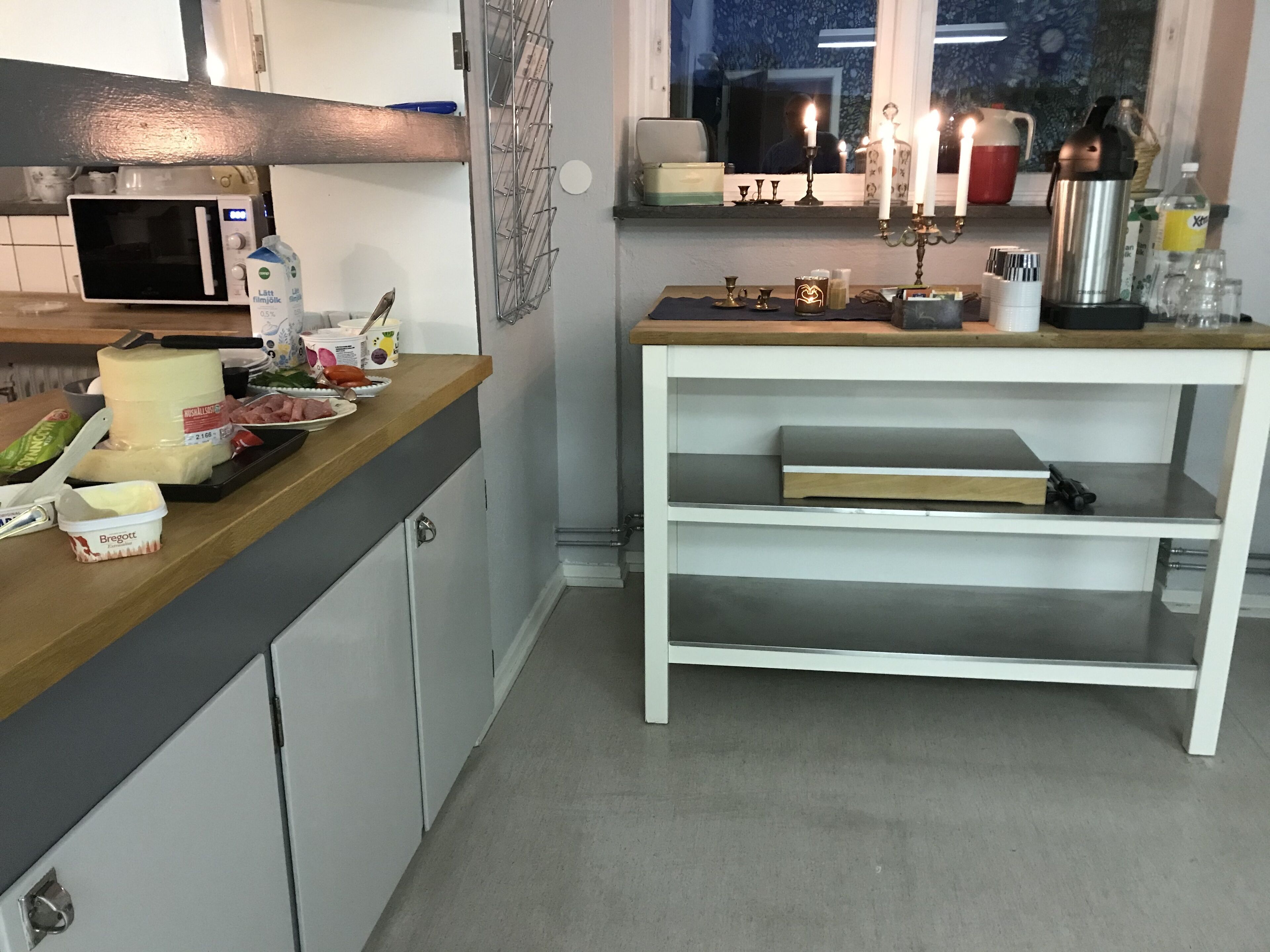 A kitchen with a microwave, a small fridge, a wooden countertop with various food items, a white shelf unit, and a window with a view of the outside.
