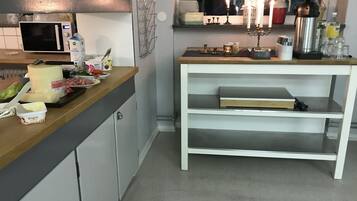A kitchen with a microwave, a small fridge, a wooden countertop with various food items, a white shelf unit, and a window with a view of the outside.
