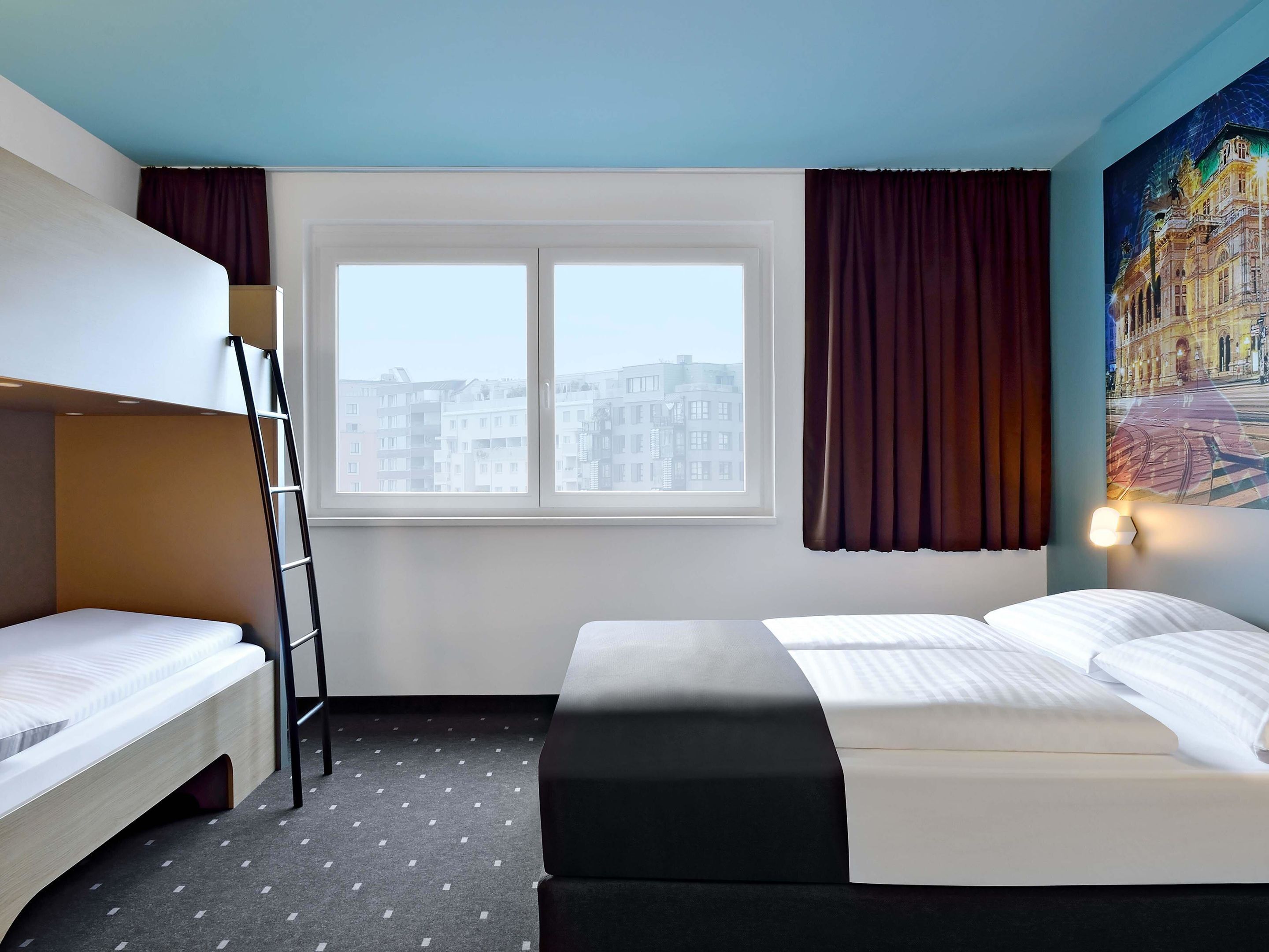 Ein Hotelzimmer mit Etagenbett, einem Einzelbett, einem Fenster mit Vorhängen, einem Bild an der Wand und einer Lampe.