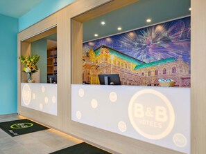 Ein Empfangsschalter mit dem B&B Hotels Logo, einer Blumenvase und einem farbenfrohen Wandbild eines Gebäudes.