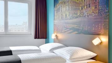 A hotel room with a bed, a window with curtains, a wall-mounted light, and a large cityscape painting.