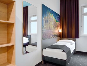 Une chambre d’hôtel avec un lit, un grand miroir, un tableau représentant un paysage urbain et une fenêtre avec des rideaux.