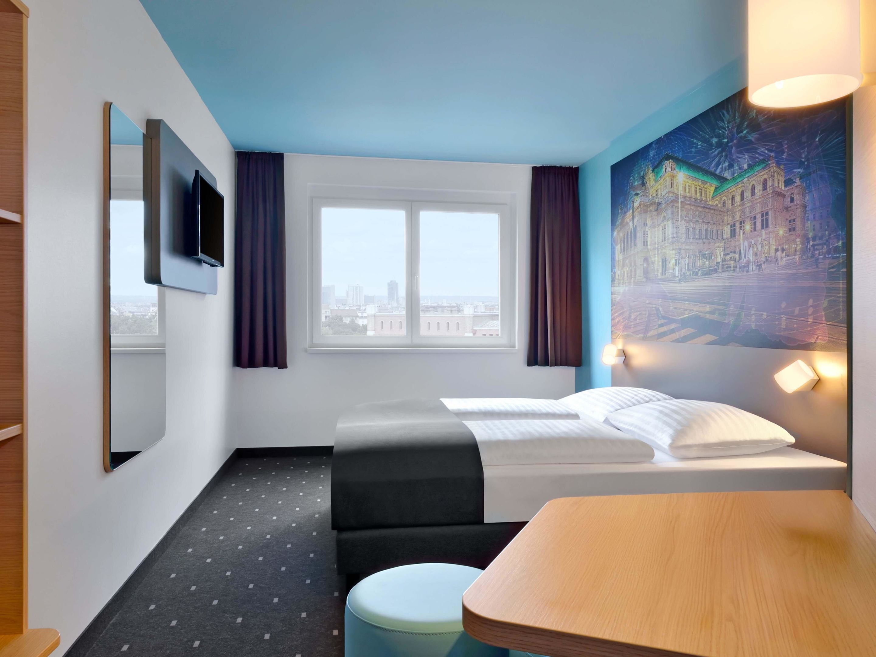 Habitación de hotel con cama, escritorio, silla, ventana con cortinas, un cuadro en la pared y televisor montado en la pared.