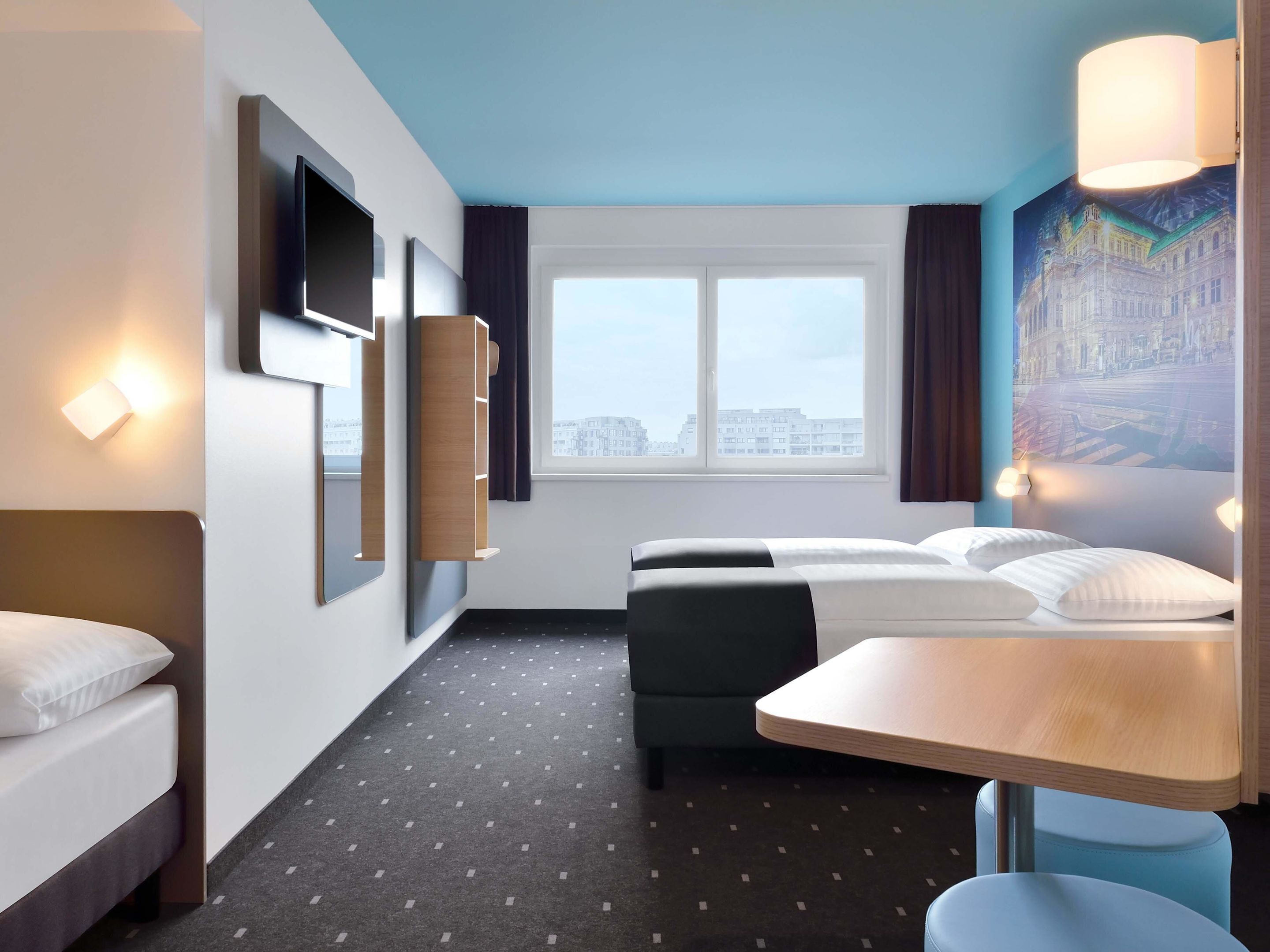 Ein modernes Hotelzimmer mit Bett, Schreibtisch, Stuhl, Fenster mit Stadtblick und Wandleuchten.