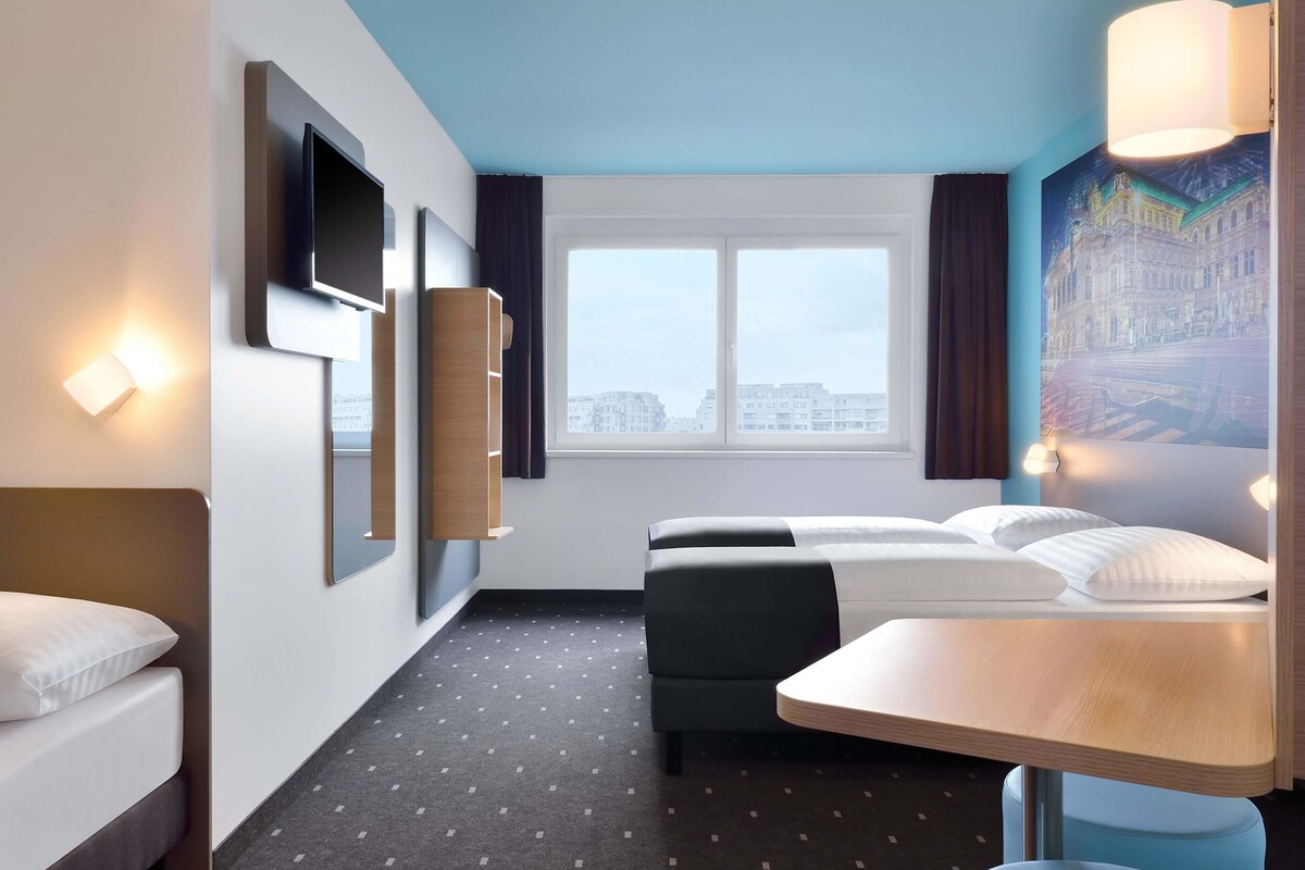 Une chambre d’hôtel moderne équipée d’un lit, d’un bureau, d’une chaise, d’une fenêtre donnant sur la ville et de lampes fixées au mur.