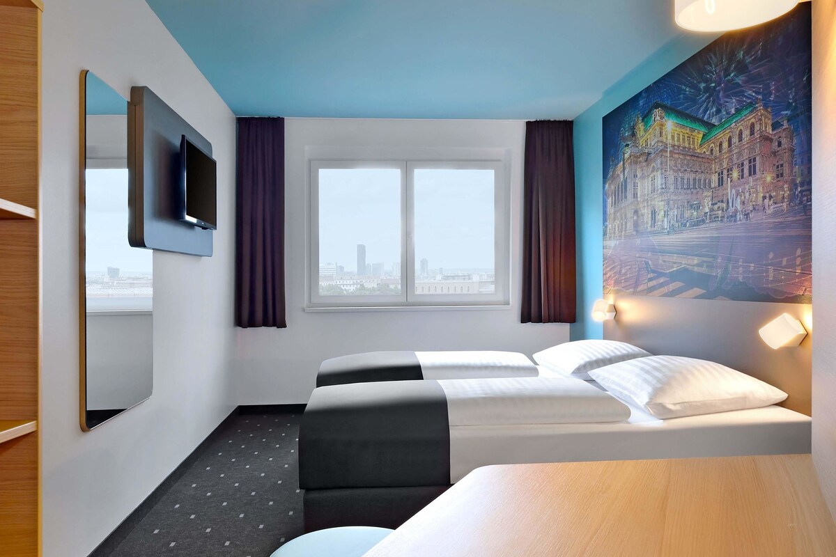 Une chambre d’hôtel avec un lit, un bureau, une chaise, une fenêtre et un tableau au mur.
