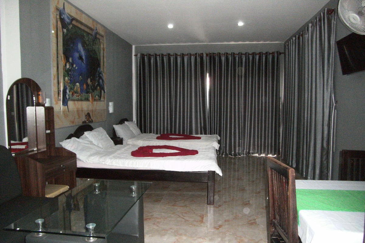 A hotel room with two beds, a glass table, a mirror, a painting, and a large window with curtains.