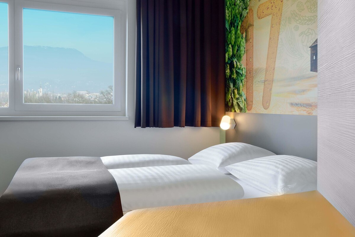 Una habitación de hotel con cama, ventana con vista a un paisaje urbano y un cuadro en la pared.