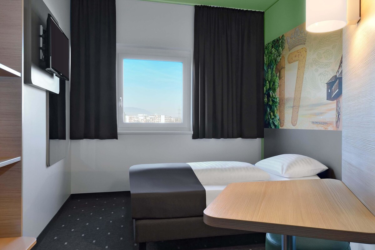 Habitación de hotel con cama, escritorio, televisor y ventana con cortinas.