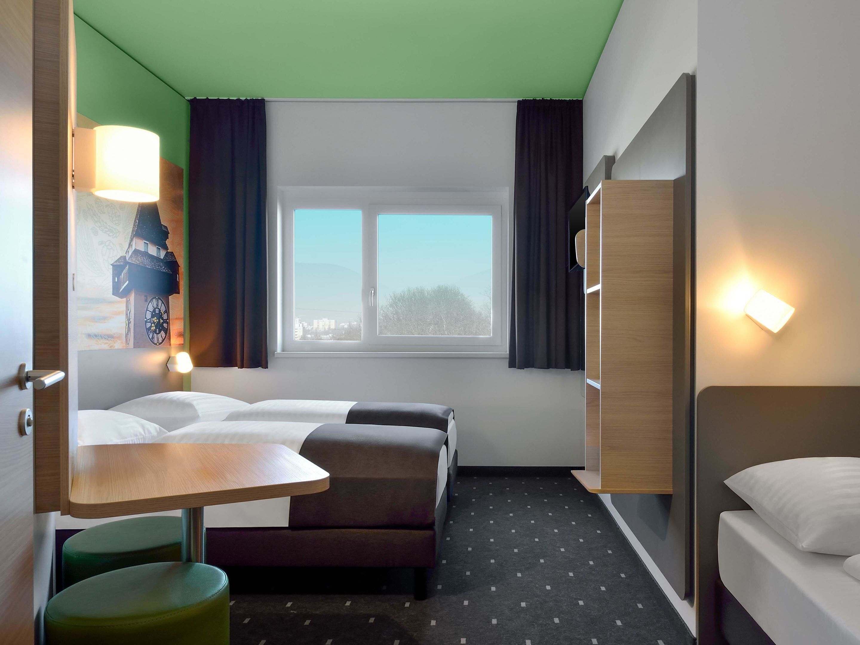 Habitación de hotel con dos camas, una mesita, una silla, una ventana con cortinas y una lámpara de pared.