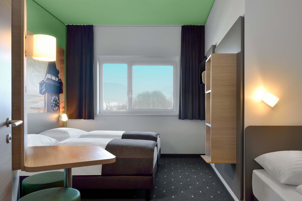 Habitación de hotel con dos camas, una mesita, una silla, una ventana con cortinas y una lámpara de pared.