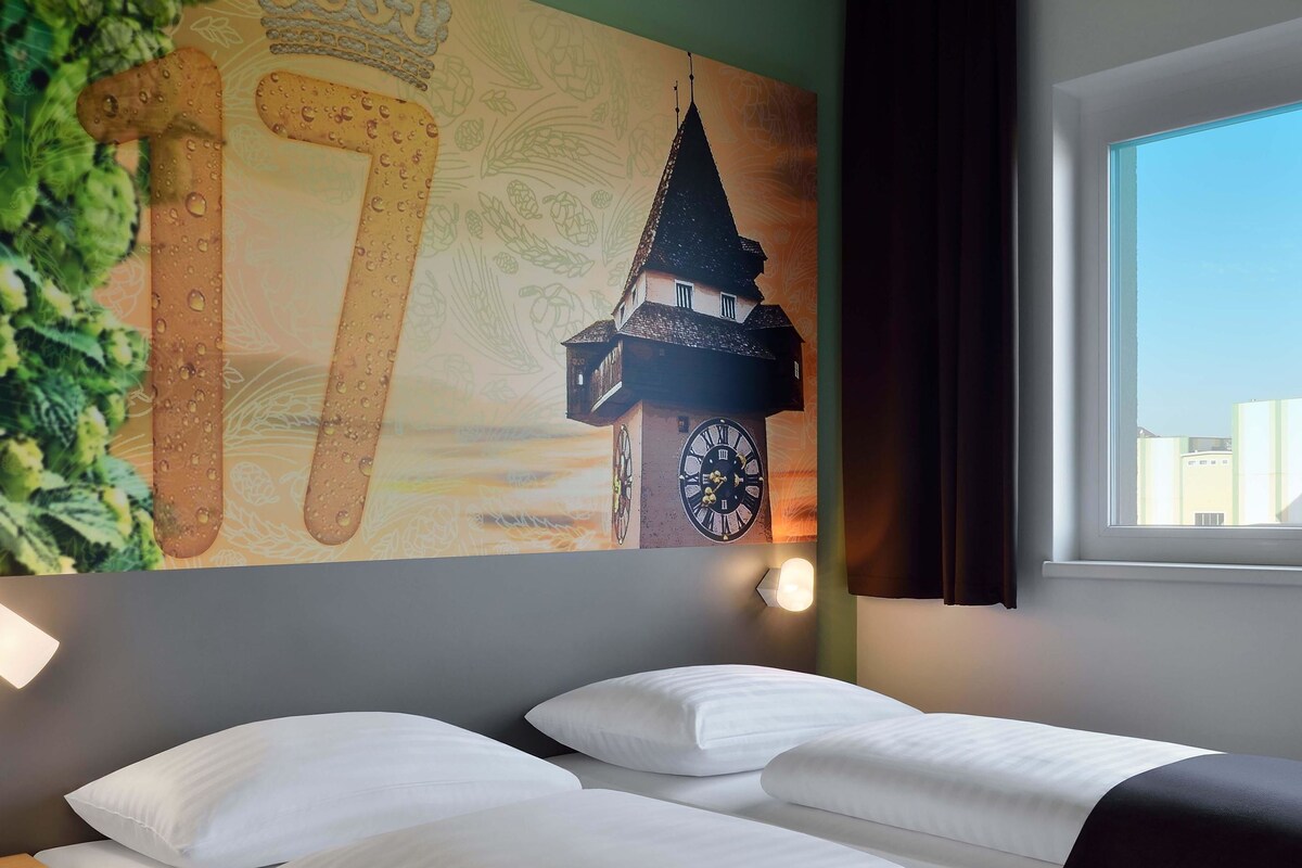 Habitación de hotel con cama, almohadas blancas, un reloj de pared y una ventana con vista a edificios.