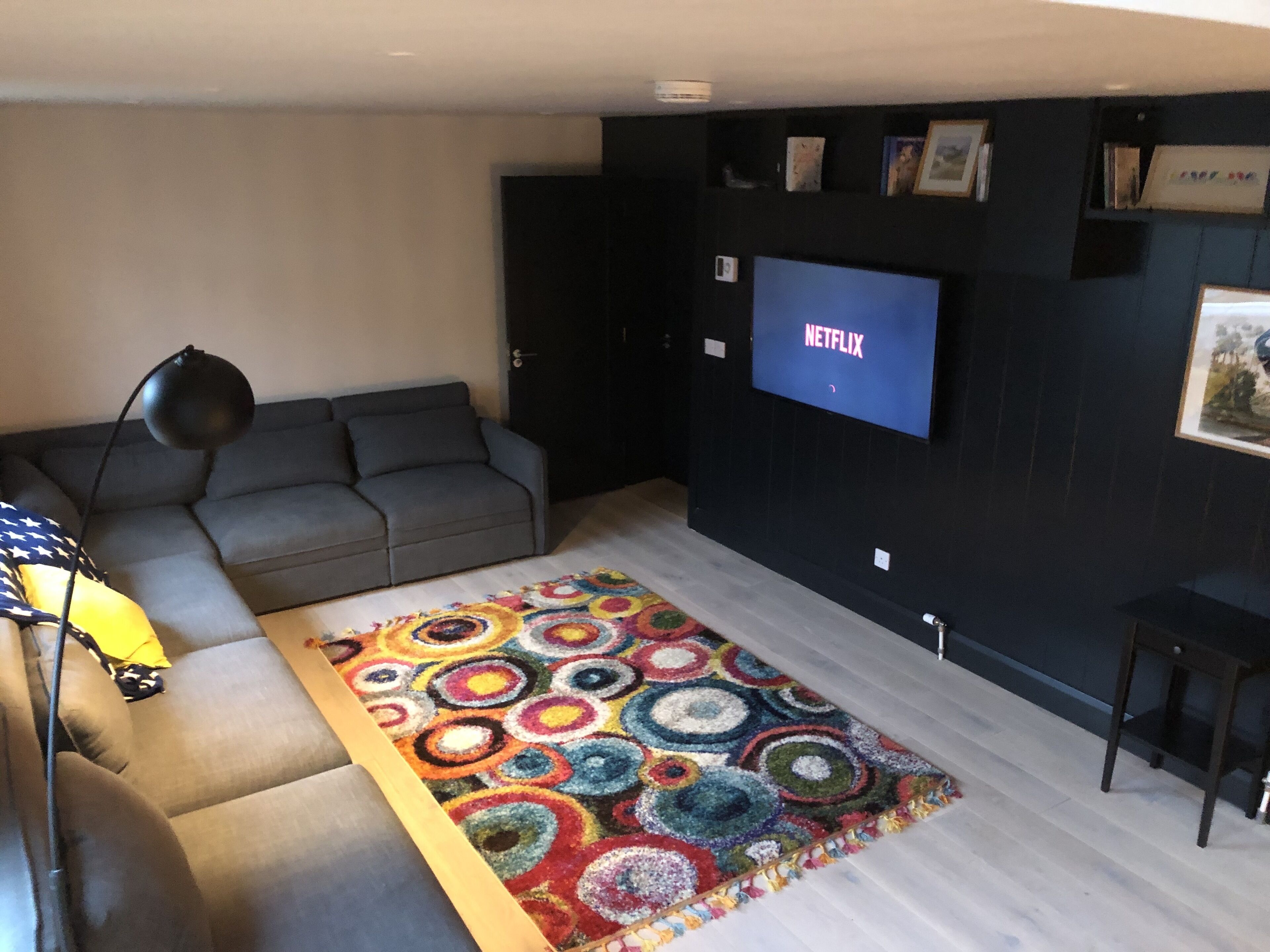 A living room with a grey sofa, a colorful circular rug, a black floor lamp, and a TV mounted on the wall displaying 'NETFLIX'.