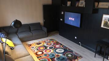 A living room with a grey sofa, a colorful circular rug, a black floor lamp, and a TV mounted on the wall displaying 'NETFLIX'.