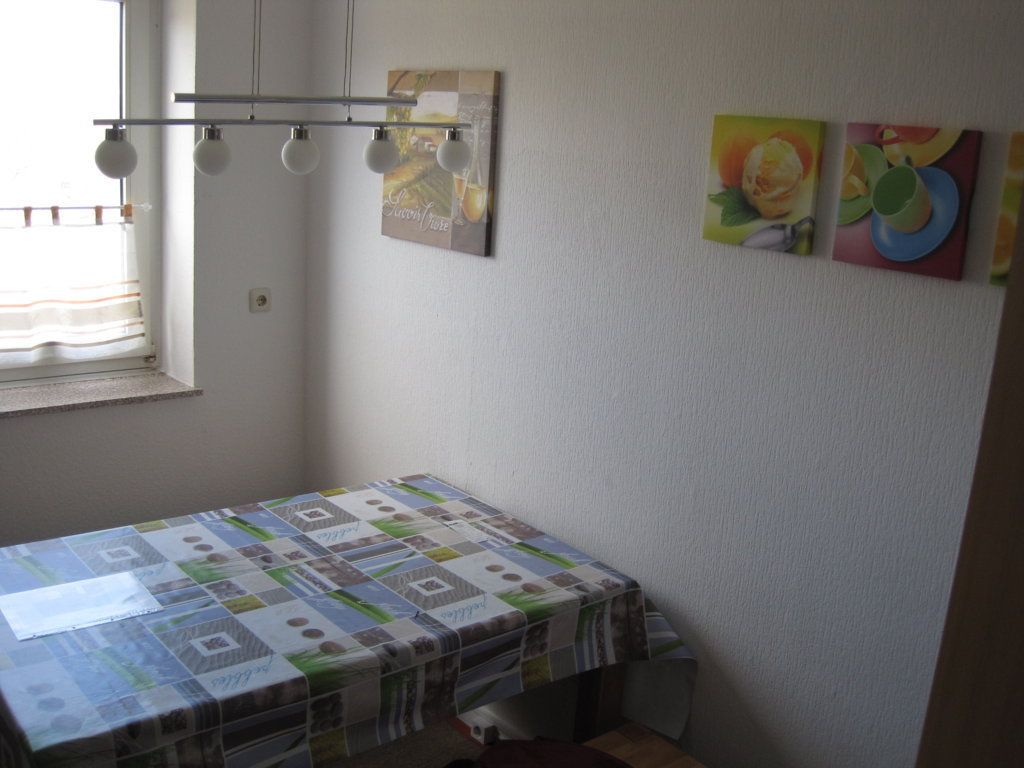 Une petite pièce avec un lit recouvert d’une courtepointe, une fenêtre avec des stores, un luminaire fixé au mur et trois tableaux encadrés.
