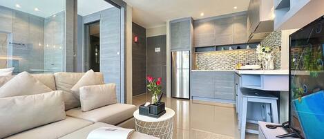 A modern living room with a sofa, a coffee table with a magazine, and a kitchen area with a refrigerator and a tiled backsplash.