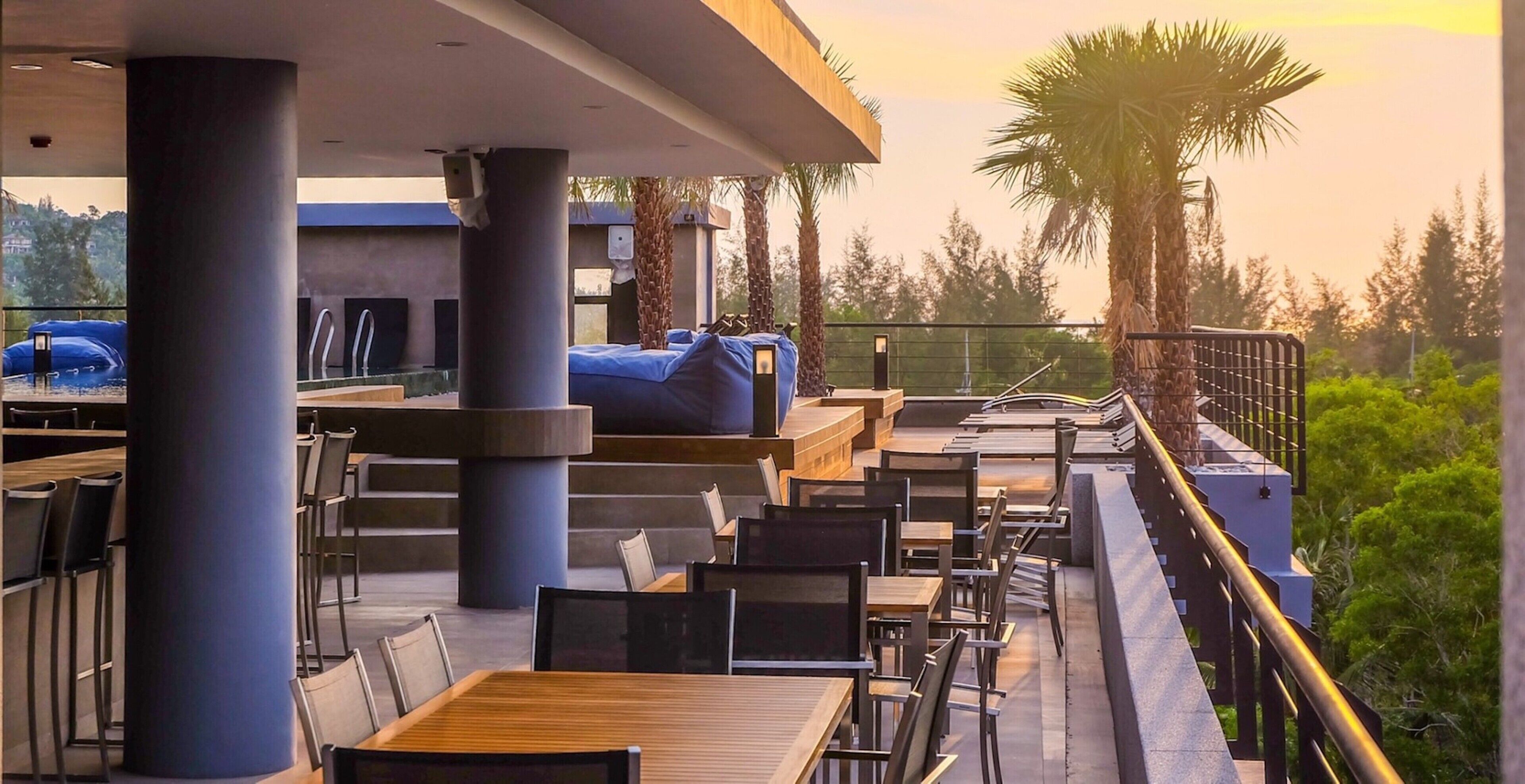 An outdoor terrace with wooden tables and chairs, a blue sofa, and a railing overlooking a garden with palm trees.