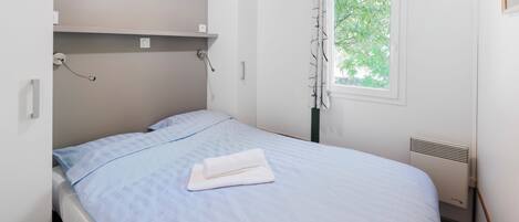 Un lit simple avec une couvre-lit bleue et des oreillers blancs, une petite étagère au-dessus du lit, une fenêtre donnant sur des arbres et un radiateur fixé au mur.