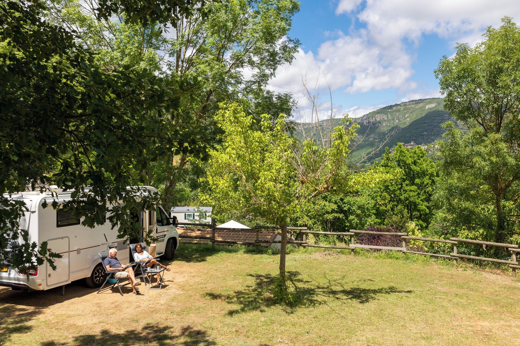 A camping site with a white RV parked on grass, surrounded by trees and a mountain view.