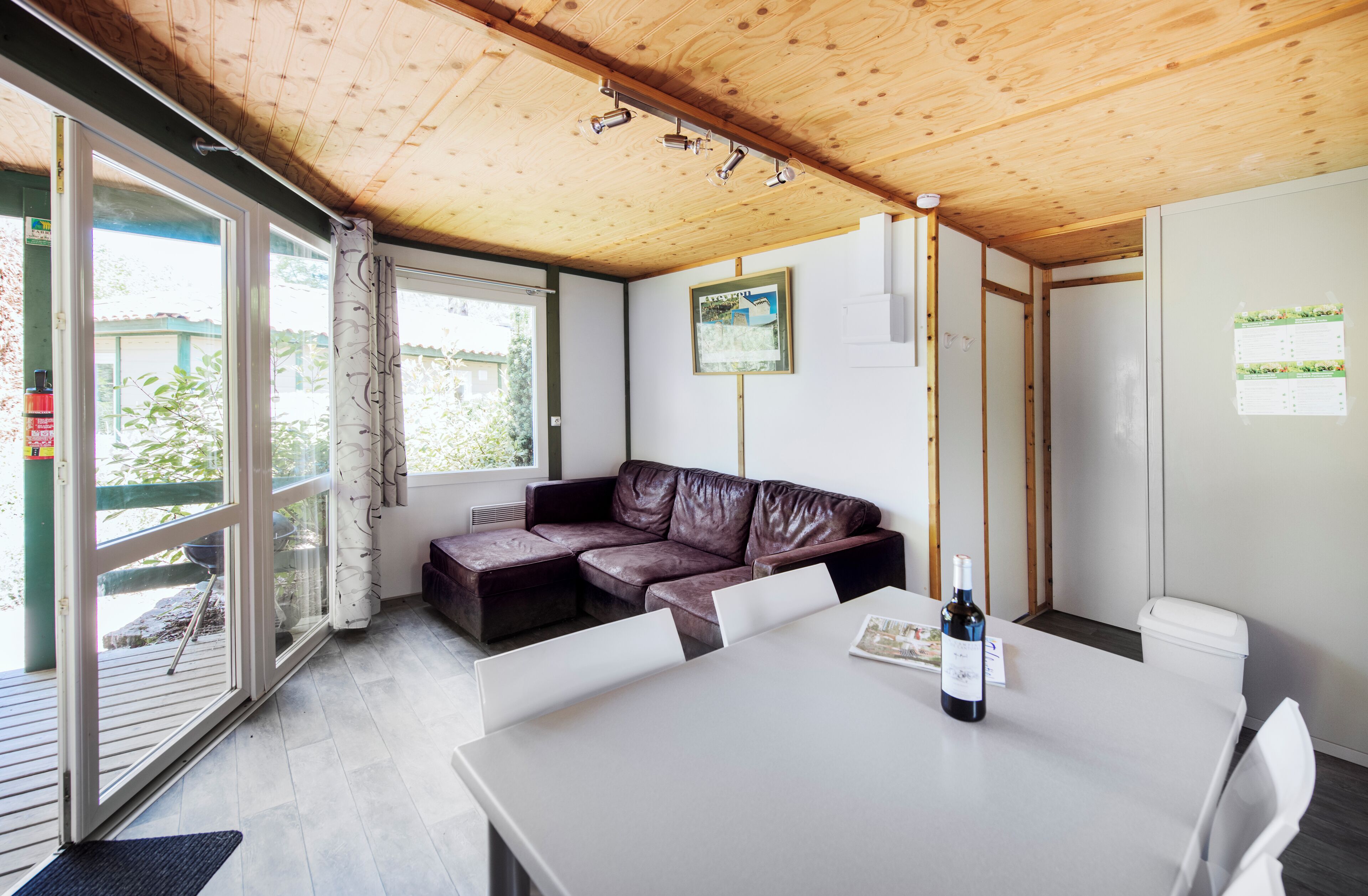 A modern room with a wooden ceiling, a leather sofa, a dining table with a bottle of wine, and large windows.