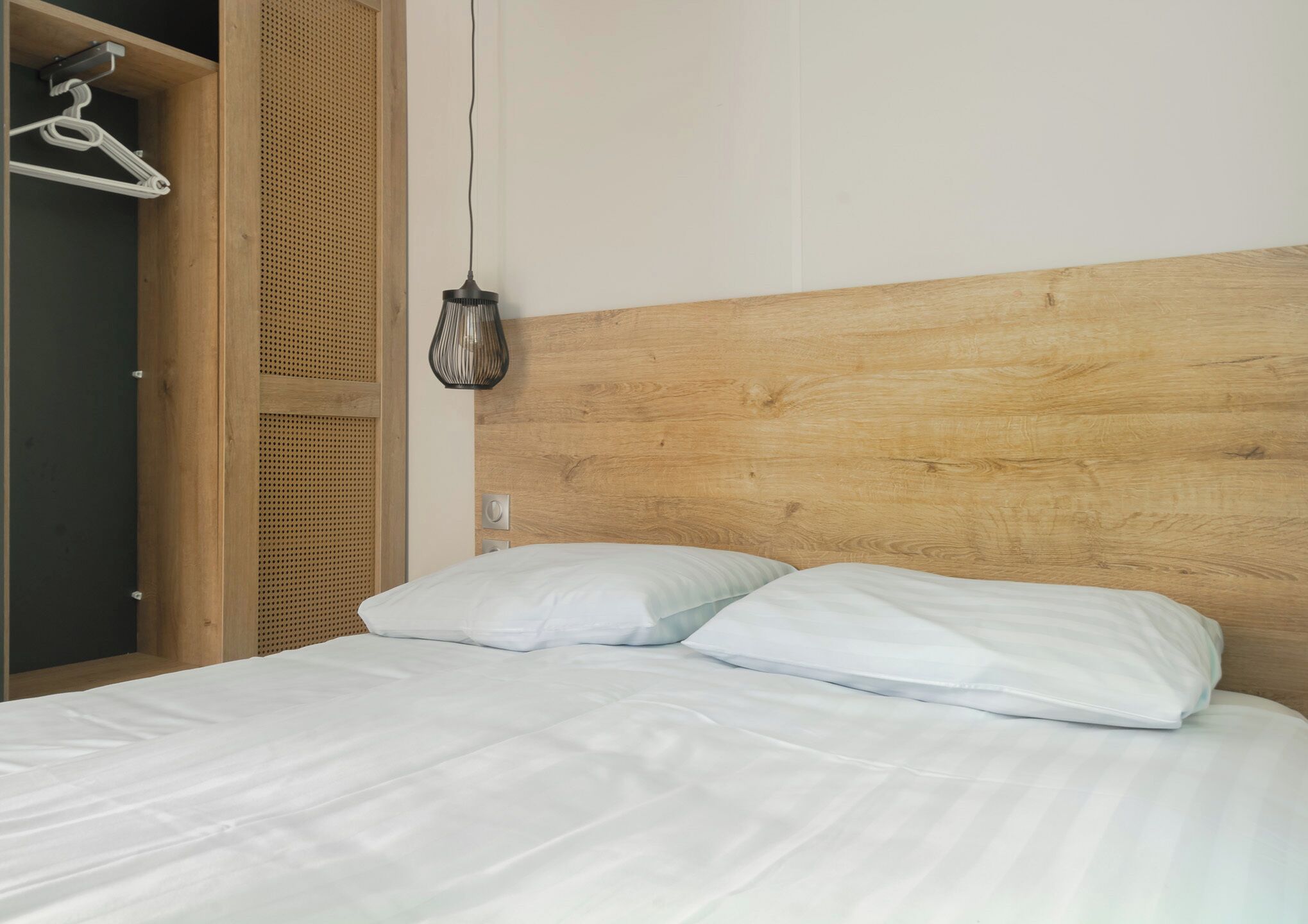 A bed with white linens, a wooden headboard, and a hanging light fixture.