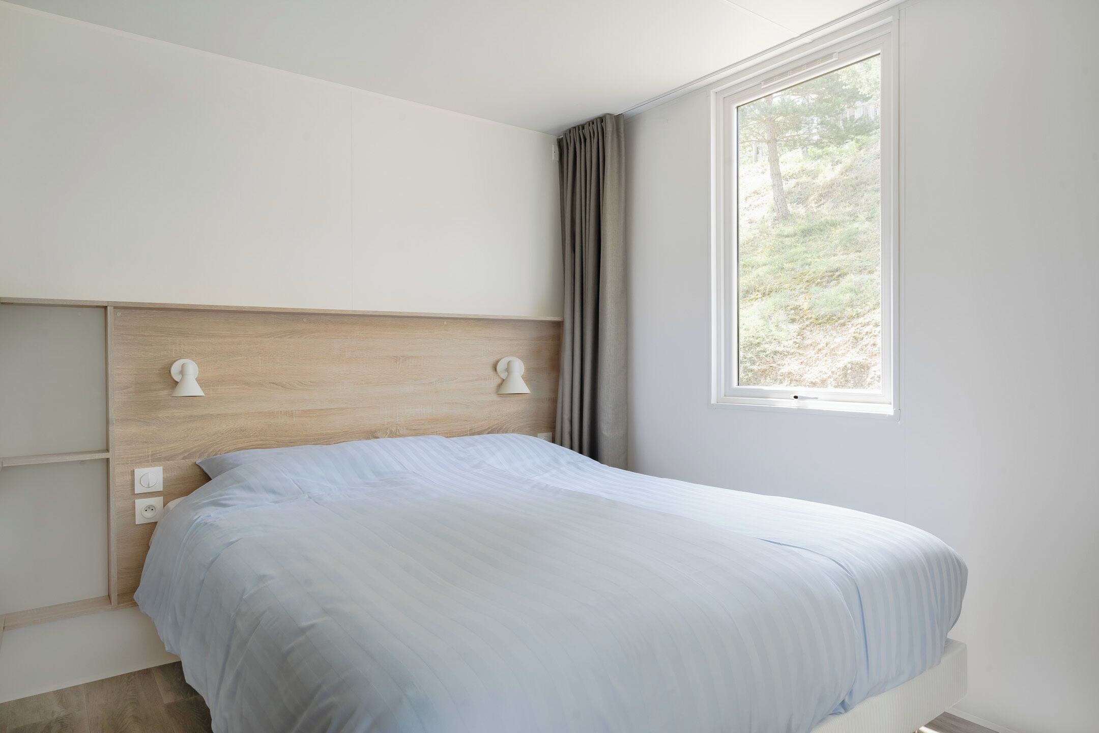 A bedroom with a wooden headboard, a bed with a light blue bedspread, a window with a view of greenery, and a white wall.