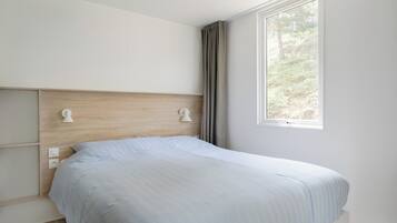 A bedroom with a wooden headboard, a bed with a light blue bedspread, a window with a view of greenery, and a white wall.
