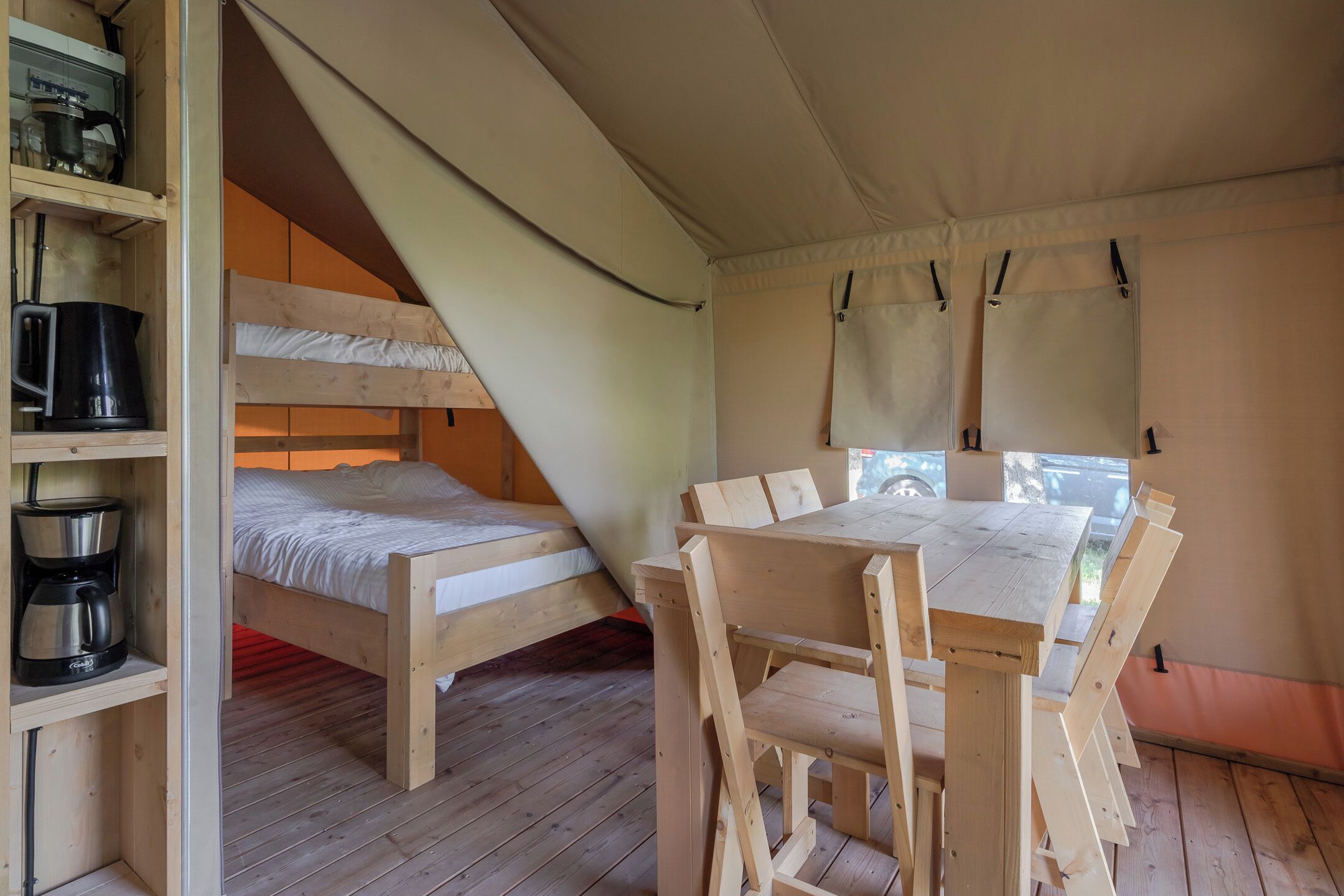 A tented accommodation with two bunk beds, a wooden table with chairs, and shelves with kitchen appliances.