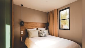 Une chambre à coucher avec une tête de lit en bois, un grand lit recouvert de linge de lit blanc, un luminaire suspendu et une fenêtre donnant sur des arbres.