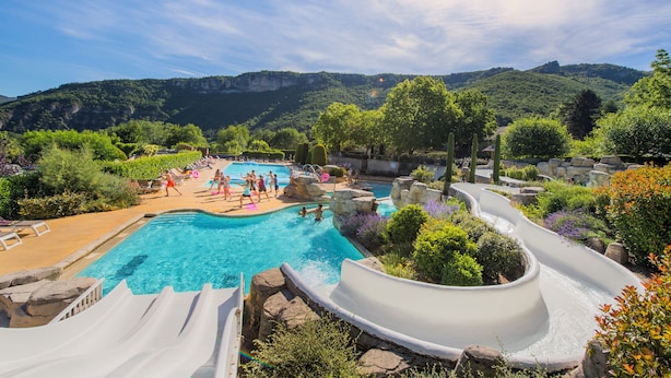 Un espace piscine doté d’un toboggan, entouré d’une végétation luxuriante et avec des montagnes en toile de fond.