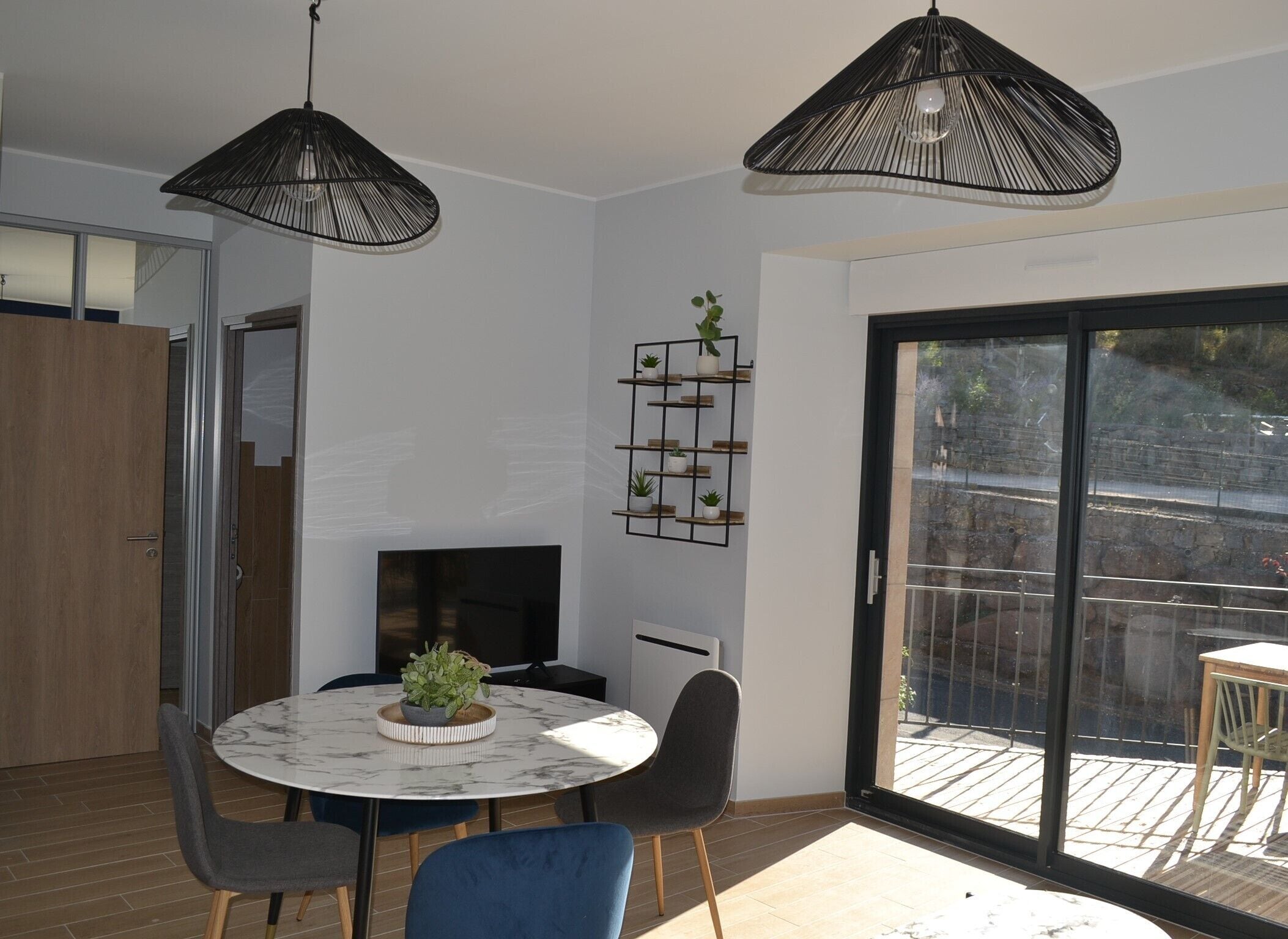 Un comedor moderno con una mesa redonda, dos sillas y una planta en el centro. Hay un televisor montado en la pared, y una estantería con plantas encima.