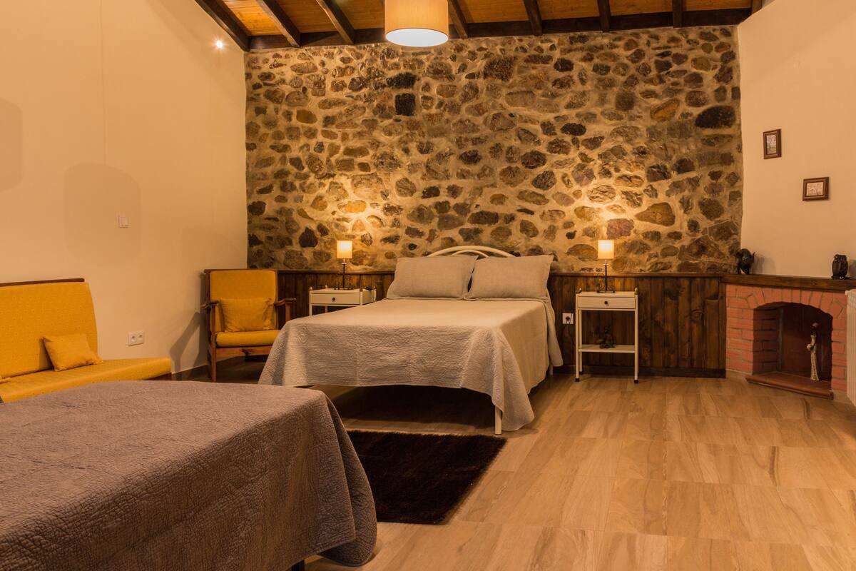 A cozy bedroom with a stone wall, a bed with a white bedspread, a wooden chair, and a small table with a lamp.