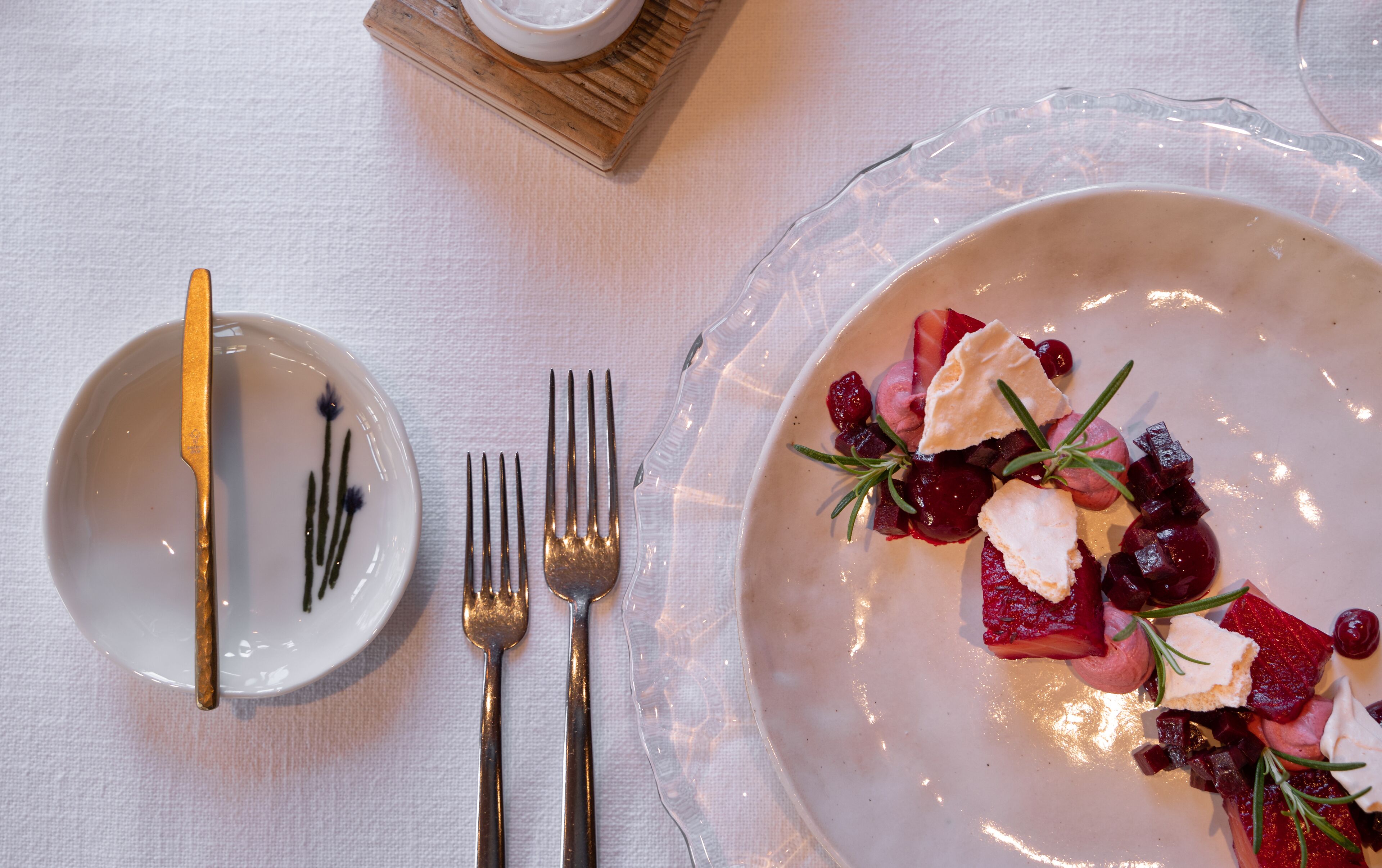 A dining table with a white tablecloth, a glass plate with a dessert, and two forks.