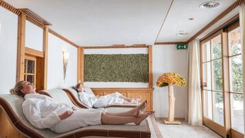 Dos camillas de spa con estructura de madera y toallas blancas, suelo de madera, un ventanal con vistas y un calentador fijado a la pared.
