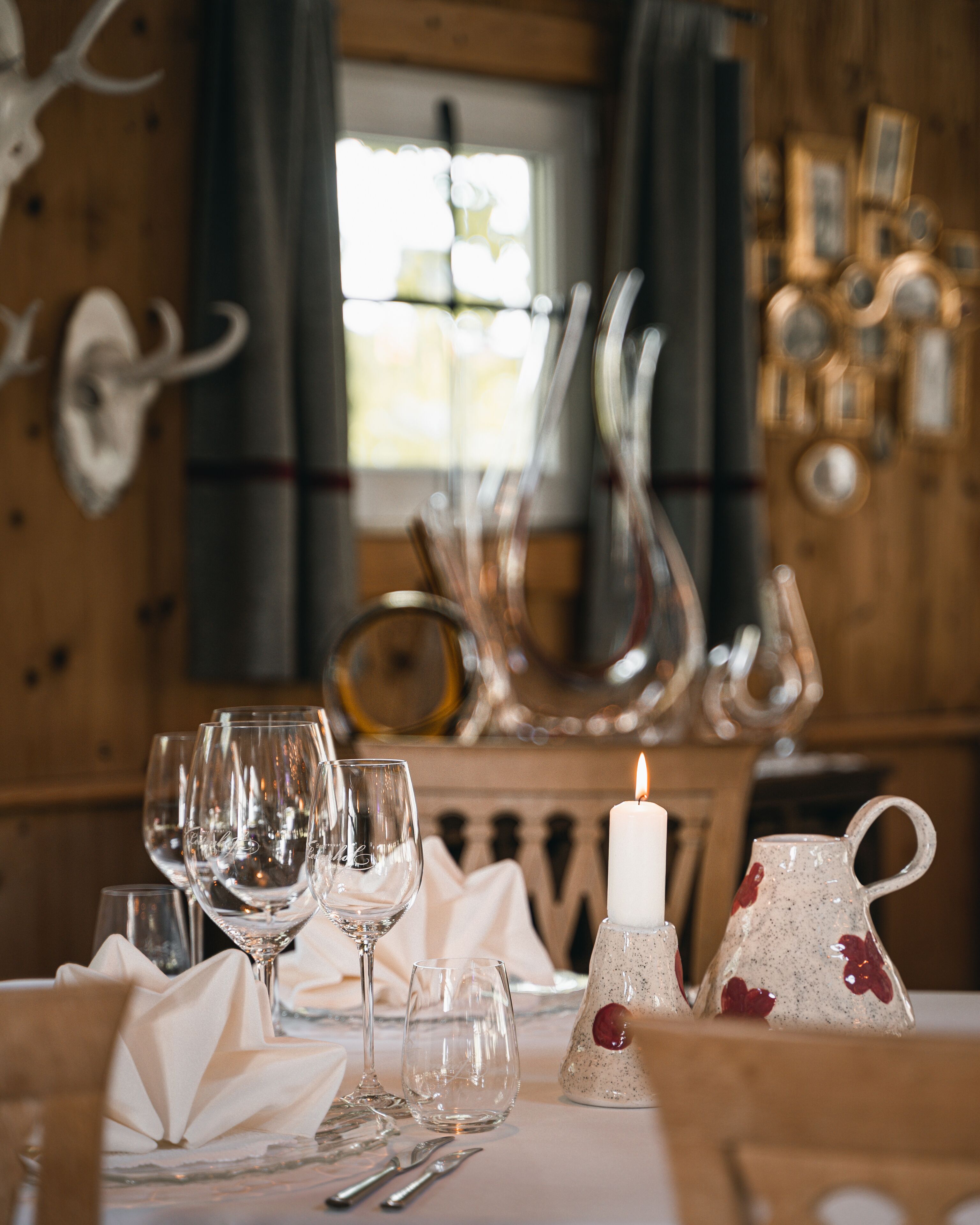 Una mesa de comedor dispuesta con copas de vino, una vela y una jarra, en una habitación con paredes de madera y una ventana.