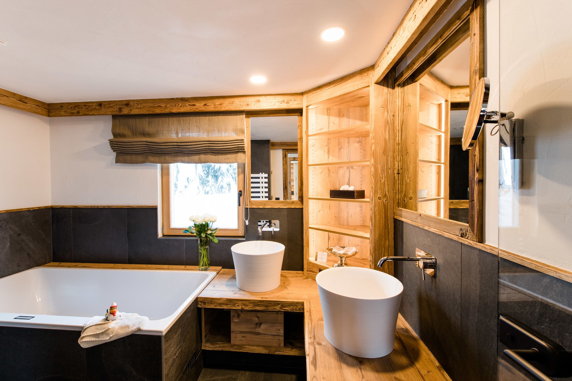 Une salle de bain moderne dotée d’une grande baignoire, de deux lavabos, d’éléments décoratifs en bois et d’une fenêtre laissant entrer la lumière naturelle.