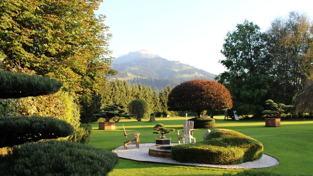 Un jardin bien entretenu, avec des haies taillées avec soin, un espace de détente circulaire agrémenté de chaises, et une montagne en arrière-plan.