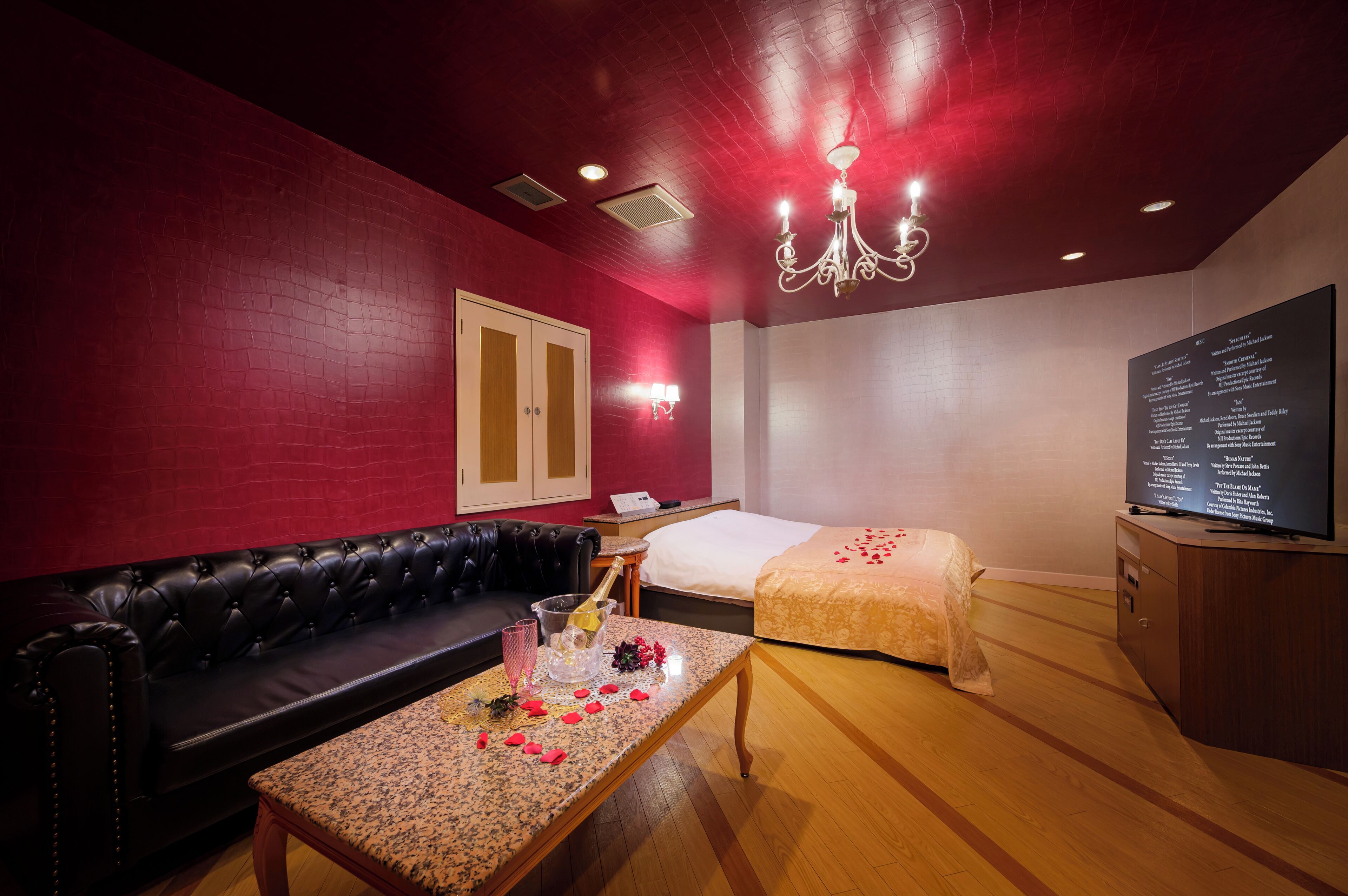 A room with a black leather sofa, a bed with a white bedspread, a small table with a bottle and glasses, and a wall with a list of music tracks.