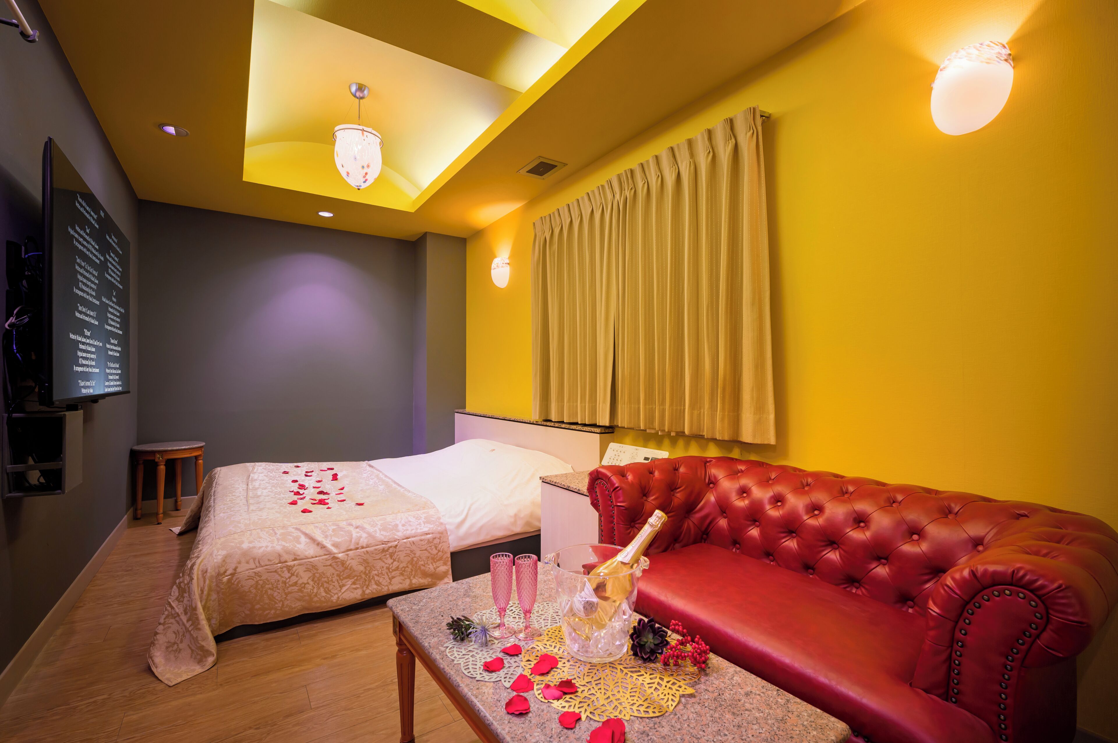 A hotel room with a bed, a red leather sofa, a small table with a bottle of champagne, and a wall-mounted TV.