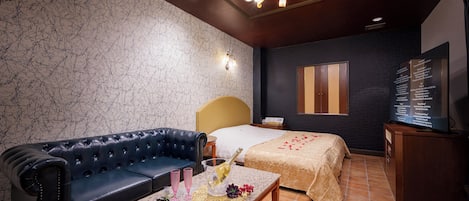Habitación de hotel con un sofá de cuero azul oscuro, una cama con colcha color beige, una mesita con una botella de champán, copas y pétalos de rosa.