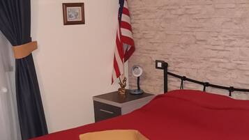 A bed with a red bedspread and a yellow pillow, a small nightstand with a lamp, an American flag, and two framed pictures on the wall.