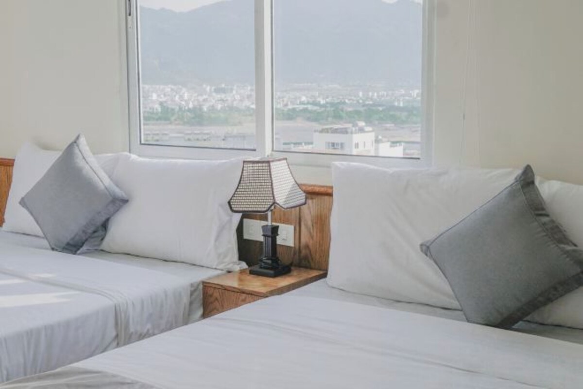 Habitación de hotel con dos camas, una mesita de noche con lámpara y un ventanal con vistas a la ciudad.